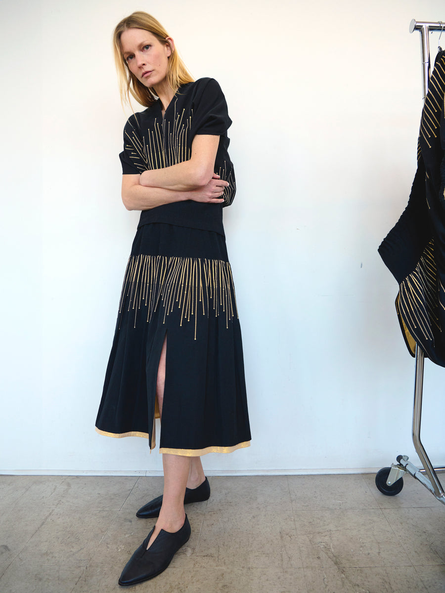 A woman with shoulder-length blonde hair stands against a white background, arms crossed. She wears the Evia Skirt from Zero + Maria Cornejo, a black dress made from recycled polyester, featuring gold vertical accents on the top and an elegant smocked skirt with a front slit. She completes the outfit with black flat shoes.