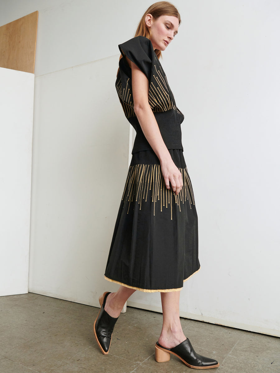 A woman with long hair is wearing a black dress adorned with vertical yellow stripes and a black belt. She is walking in a minimalistic room with grey flooring and white walls, completing her look with black open-back heeled mules that rival the elegance of the Evia Skirt by Zero + Maria Cornejo.