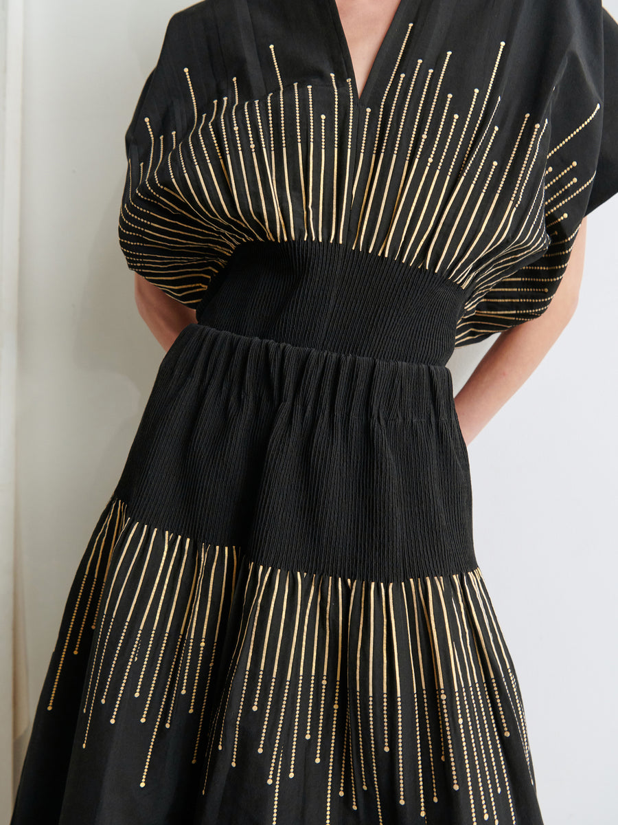 A person stands against a plain, light-colored background, dressed in the Evia Skirt by Zero + Maria Cornejo. The black dress is beautifully adorned with vertical golden stripes across the chest and waist. It features organic metallic smocking and a v-neck, cinched at the waist to create a flared fil coupe woven skirt.