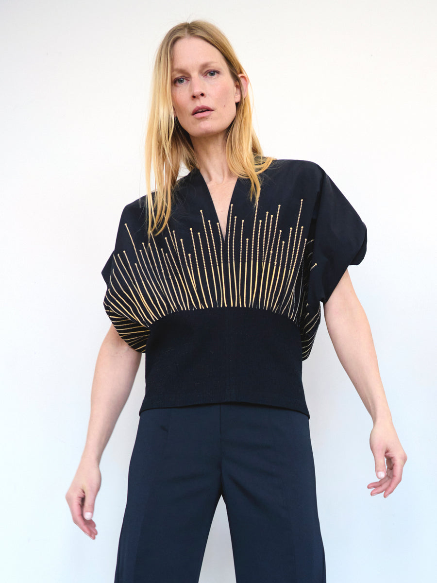 A woman with long blonde hair stands against a plain background. She is wearing the Eva Top by Zero + Maria Cornejo, featuring a Fil coupe v-neck design in navy blue adorned with a geometric gold pattern emanating from the midsection, paired with matching navy blue pants. Her arms are slightly outstretched, and she has a neutral expression on her face.