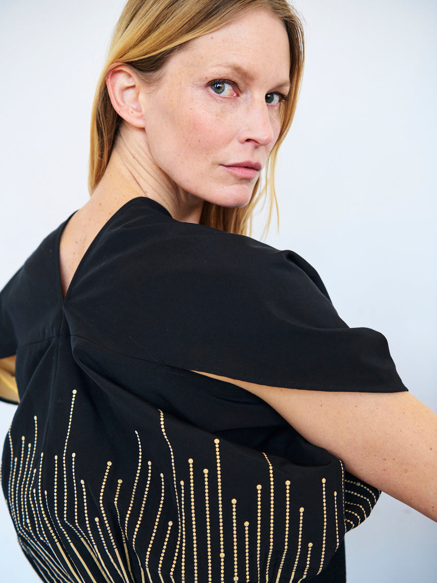 A person with long blonde hair and fair skin looks over their right shoulder, wearing the Eva Top by Zero + Maria Cornejo, which is a black garment adorned with gold bead or sequin detailing. The plain, light-colored background draws focus to the individual and their attire, which features GRS certified polyester for eco-conscious elegance.