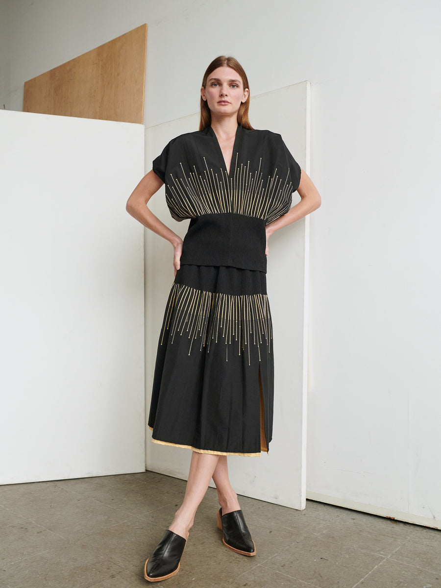 A woman stands with her hands on her hips, wearing an Eva Top by Zero + Maria Cornejo, which features a black design with metallic sparkler details and a smocked waist. The top showcases gold vertical line patterns throughout. She has long brown hair and is standing in a minimalist room with white and wooden panels in the background, complemented by her black slip-on shoes.