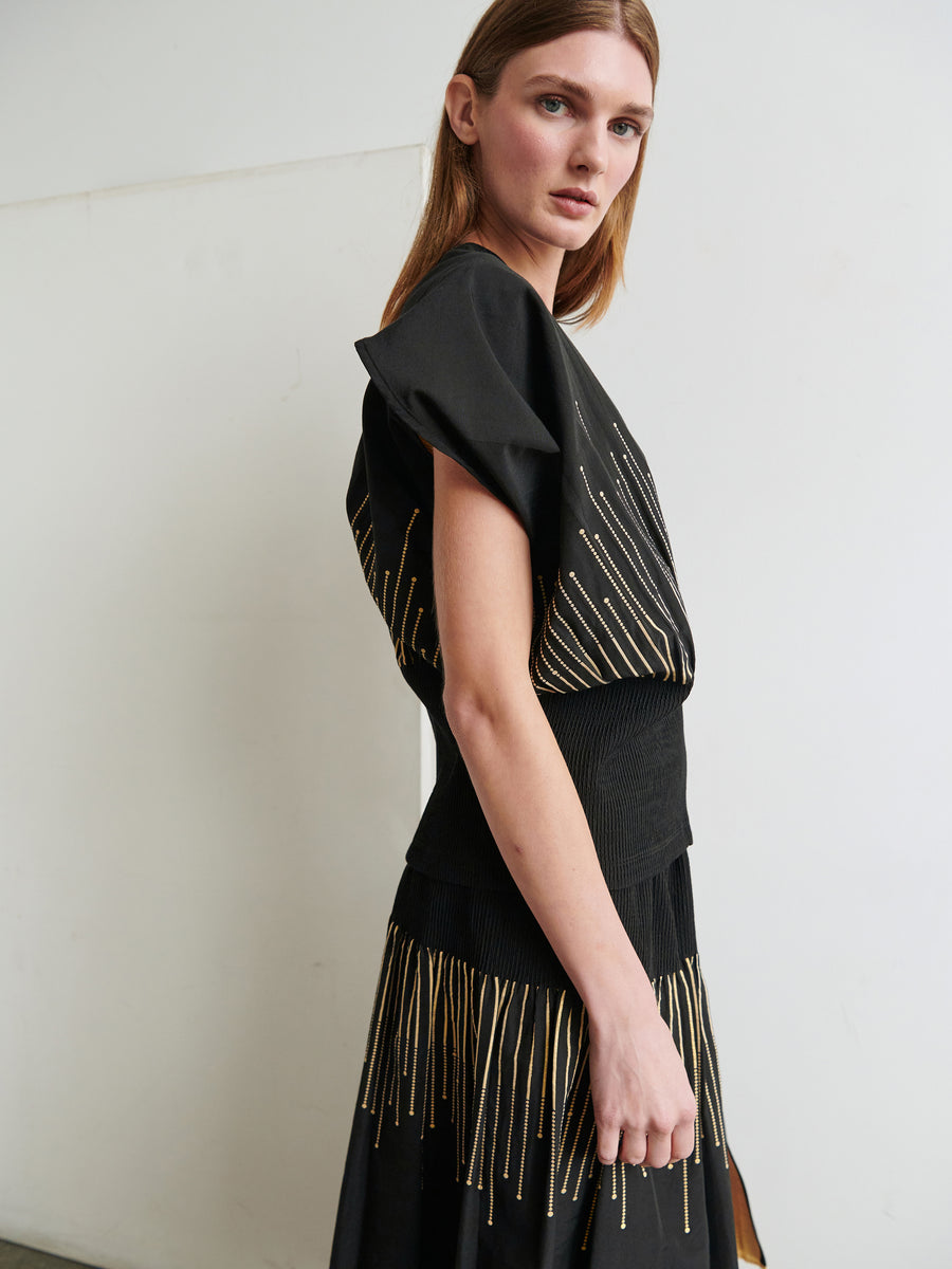 A woman stands indoors, showcasing the Eva Top by Zero + Maria Cornejo. The top features black fabric with gold vertical stripes and a subtle metallic sparkler design. She is turned slightly to the side, looking towards the camera with a neutral expression. The background is simple and white.