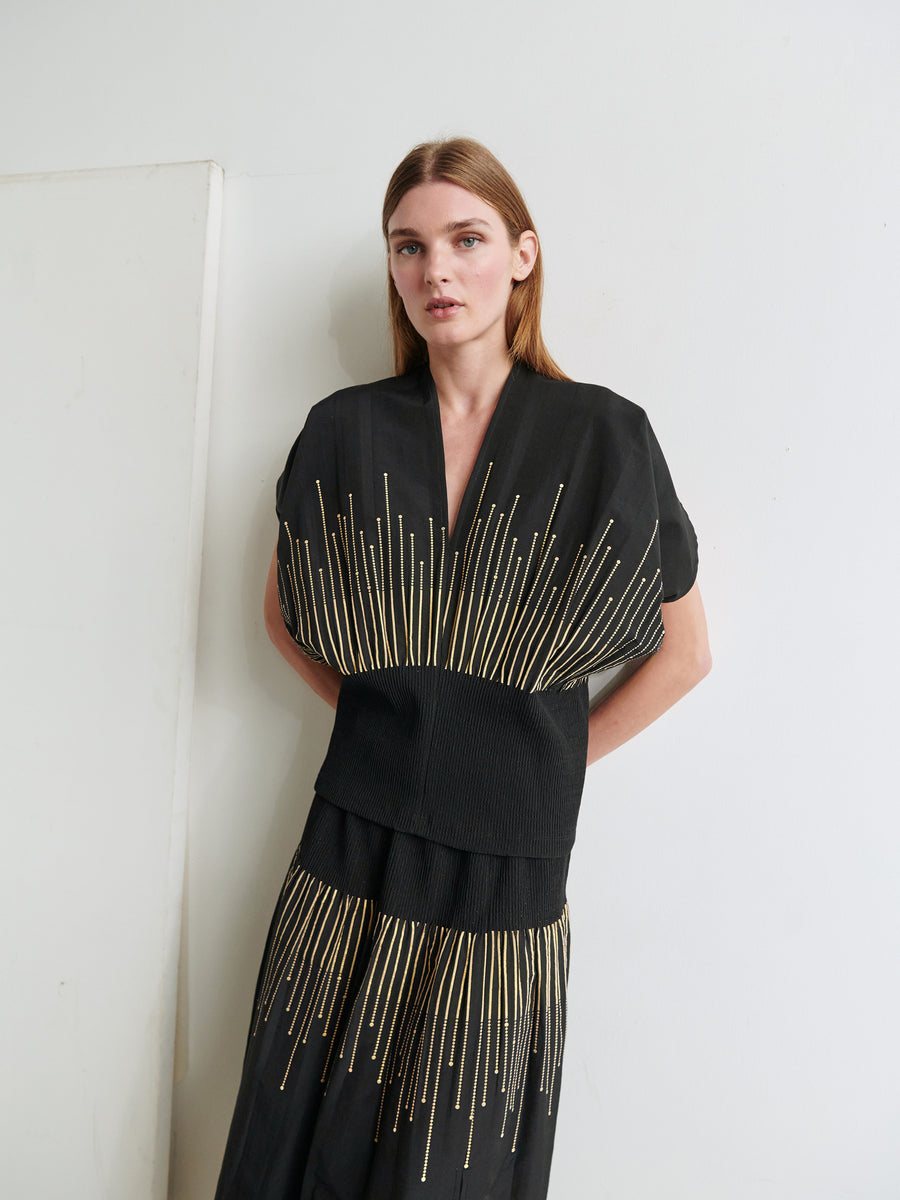 A person with long, straight hair stands against a white wall, wearing the black Eva Top from Zero + Maria Cornejo. The outfit features intricate gold linear details emanating from the waist and chest, creating a striking pattern. The person looks directly at the camera with a calm expression.