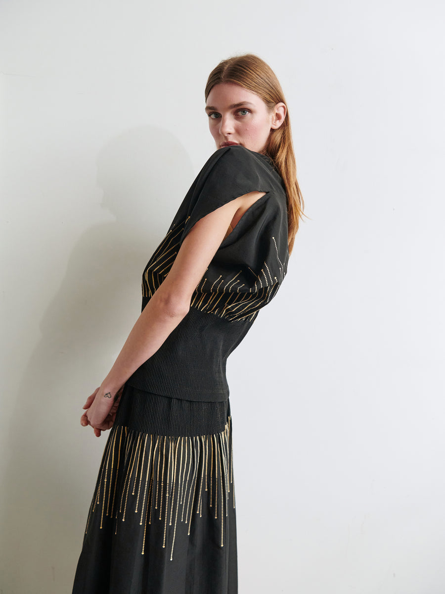 A young woman with long, straight brown hair stands against a plain white wall, turned slightly to the side. She is wearing the Eva Top by Zero + Maria Cornejo, a black outfit featuring gold vertical line patterns and a smocked waist. She gazes directly at the camera with a neutral expression.