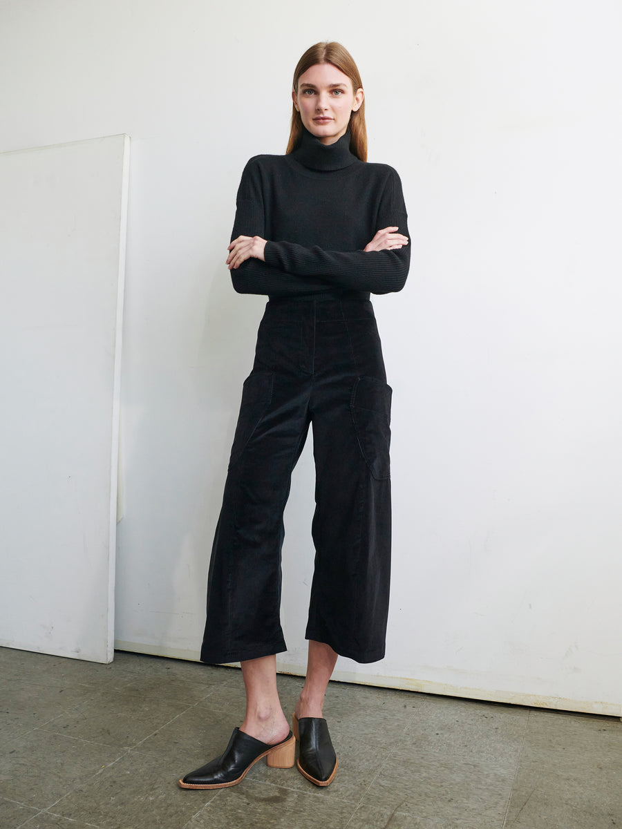 A woman stands against a plain white background with her arms crossed. She is wearing a black turtleneck sweater, the Curved Eda Culotte by Zero + Maria Cornejo, and black mule shoes with a wooden heel. The floor she stands on is grey.