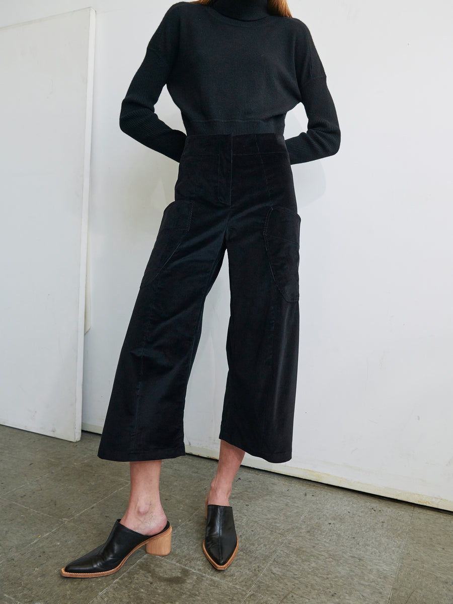 A person stands indoors against a plain backdrop, wearing a black turtleneck sweater, Curved Eda Culotte from Zero + Maria Cornejo featuring front patch pockets and wide legs, and black heeled loafers. The individual's hands are placed at their hips, emphasizing the outfit's sleek and modern design.