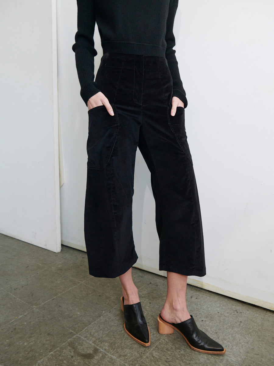 A person is standing against a white wall, wearing a black long-sleeve sweater, black Curved Eda Culotte pants by Zero + Maria Cornejo, and black heeled mules. Their hands are in their pockets, and the outfit is stylish and monochromatic. The floor is gray and appears to be concrete.