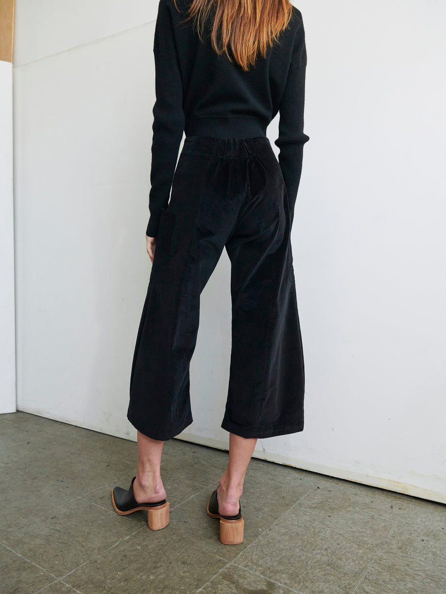 A person with long, light brown hair is standing with their back to the camera, dressed in a black long-sleeved top and the Curved Eda Culotte by Zero + Maria Cornejo, which features wide-leg corduroy cropped design and front patch pockets. They are in a room with a light-colored wall and gray floor, completing their ensemble with black heeled mules.