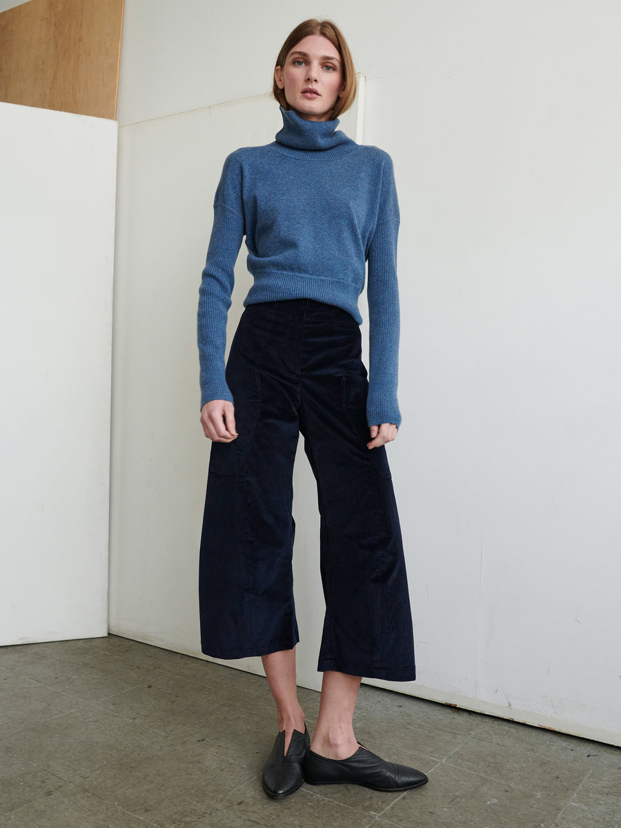 A person stands against a plain wall, wearing a blue turtleneck sweater and Curved Eda Culotte by Zero + Maria Cornejo. They have on black slip-on shoes and are posed with a relaxed demeanor, looking directly at the camera. The setting appears minimalist and indoors.