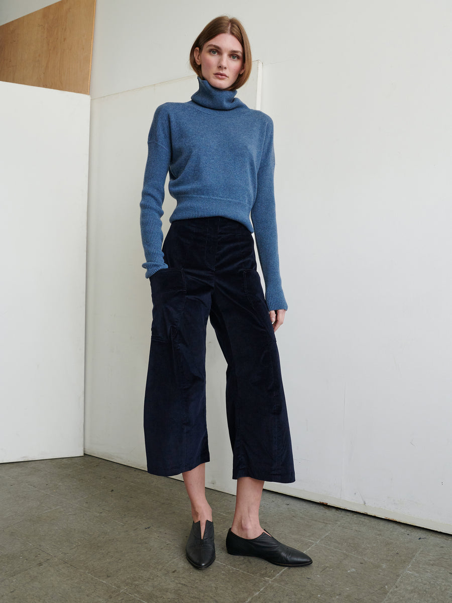 A person stands indoors against a white wall, dressed in a blue turtleneck sweater and the Curved Eda Culotte from Zero + Maria Cornejo. They wear black loafers and have straight, shoulder-length hair. Their hands are by their sides, and they gaze directly at the camera with a neutral expression.