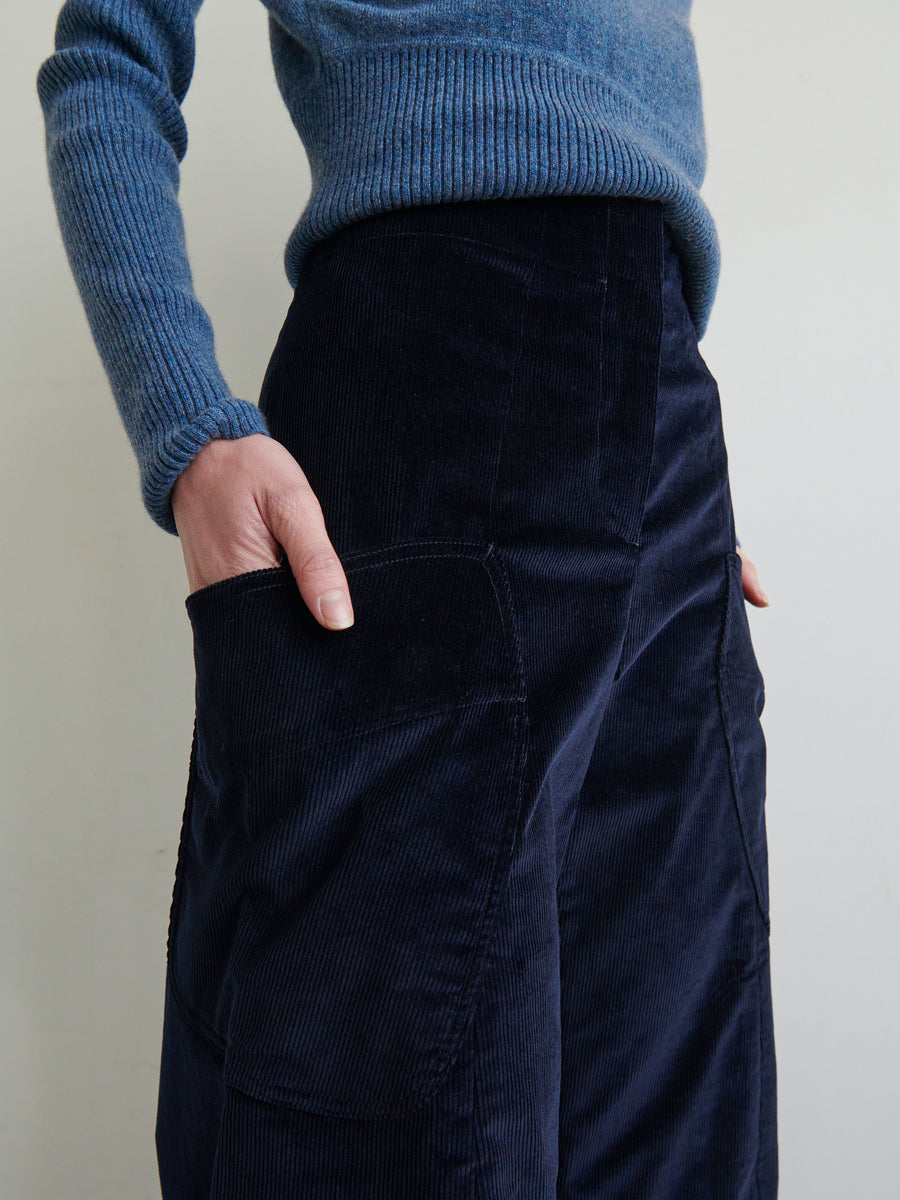 A person is shown from the chest down, wearing a blue ribbed sweater and the Curved Eda Culotte by Zero + Maria Cornejo in dark blue. Their left hand is tucked casually into one of the front patch pockets. The background is plain and neutral.