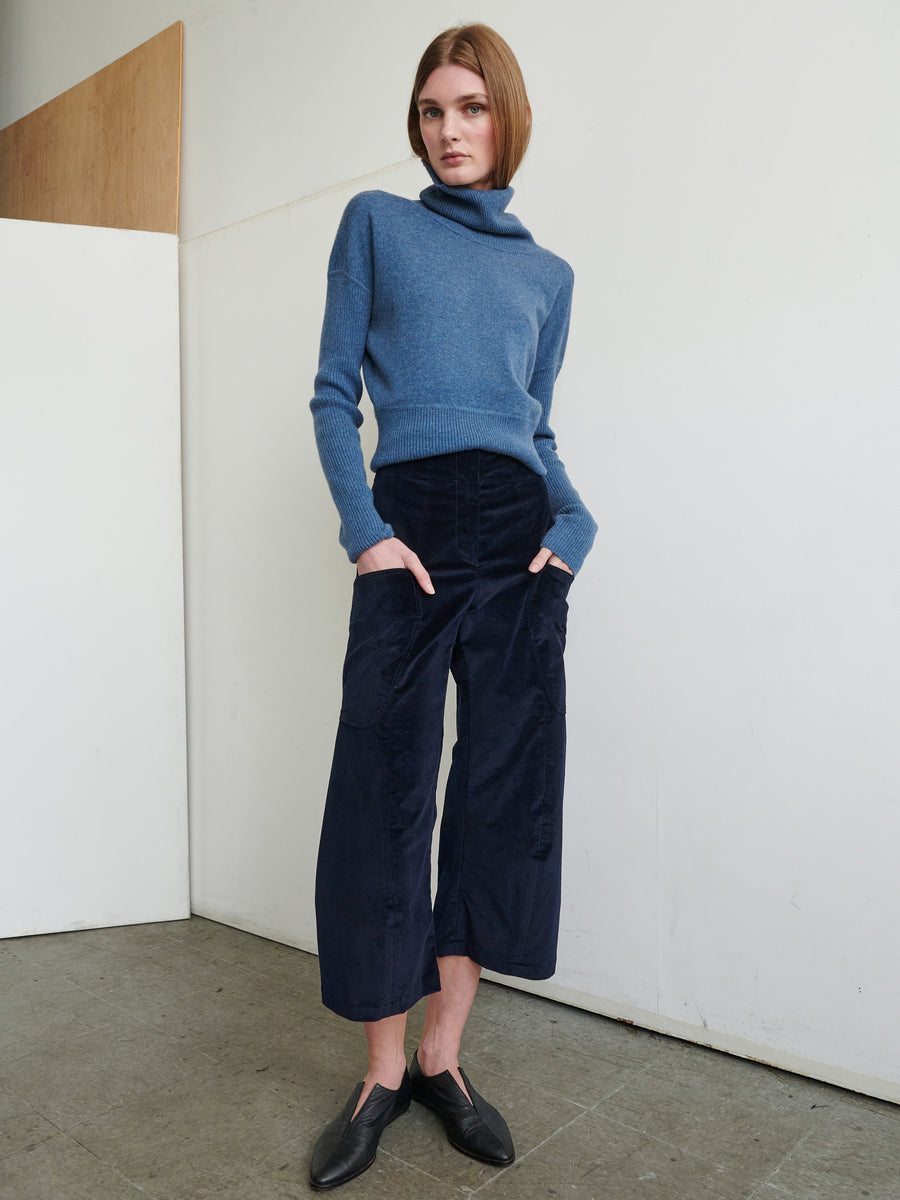 A person with short, straight hair stands against a white wall. They wear a blue sweater with a high collar, the Curved Eda Culotte dark blue corduroy cropped pants from Zero + Maria Cornejo, and black shoes. Their hands are in their front patch pockets, and they gaze directly at the camera.