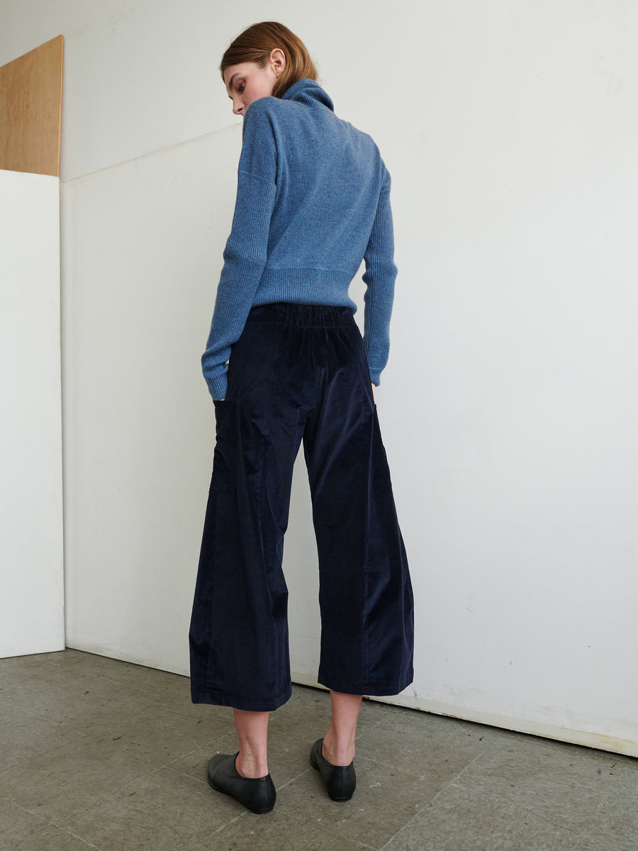 A person with short hair stands facing a white wall, wearing a light blue turtleneck sweater with front patch pockets, navy blue Curved Eda Culottes by Zero + Maria Cornejo, and black flat shoes. The person has their hands in the sweater pockets. The setting appears to be indoors with concrete flooring.