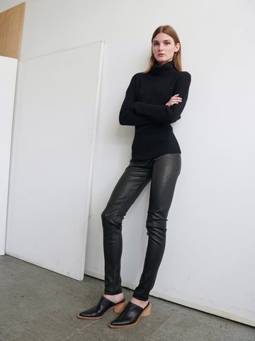 A person with long hair stands casually against a wall with arms crossed, wearing a black turtleneck sweater, the Leather One Seam Pant by Zero + Maria Cornejo featuring an elastic waistband, and black slip-on shoes with a low wooden heel. The background includes a blank canvas propped against the wall.