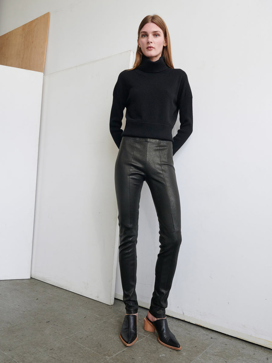 A person with long hair is leaning against a wall in a minimalist room. They are wearing a black turtleneck sweater, Leather One Seam Pant by Zero + Maria Cornejo, and black slip-on shoes. The floor is gray, and a wooden panel is partially visible on the wall.