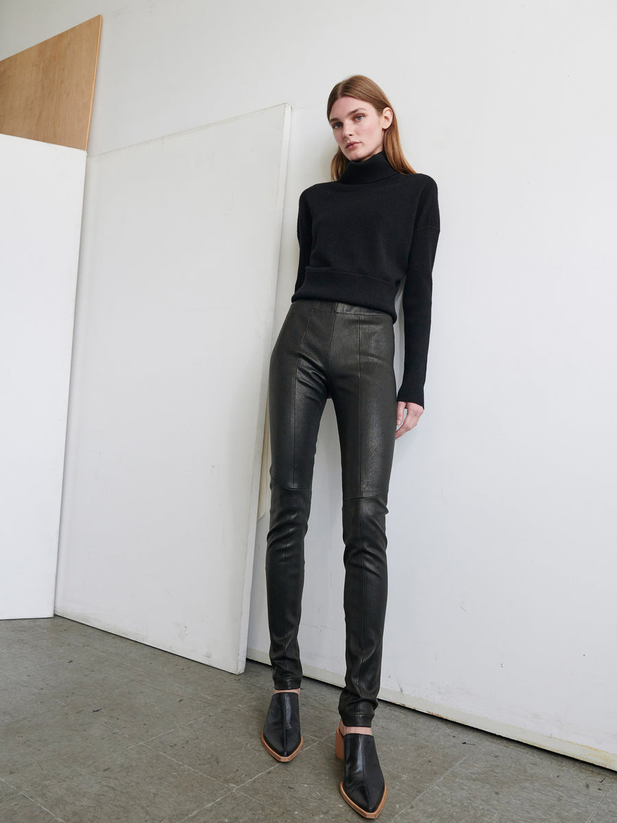 In a minimalist room, a person with long hair stands against a white wall, wearing a black turtleneck sweater paired with the Leather One Seam Pant from Zero + Maria Cornejo. They complete the outfit with black pointed shoes featuring brown soles and look ahead with a neutral expression.