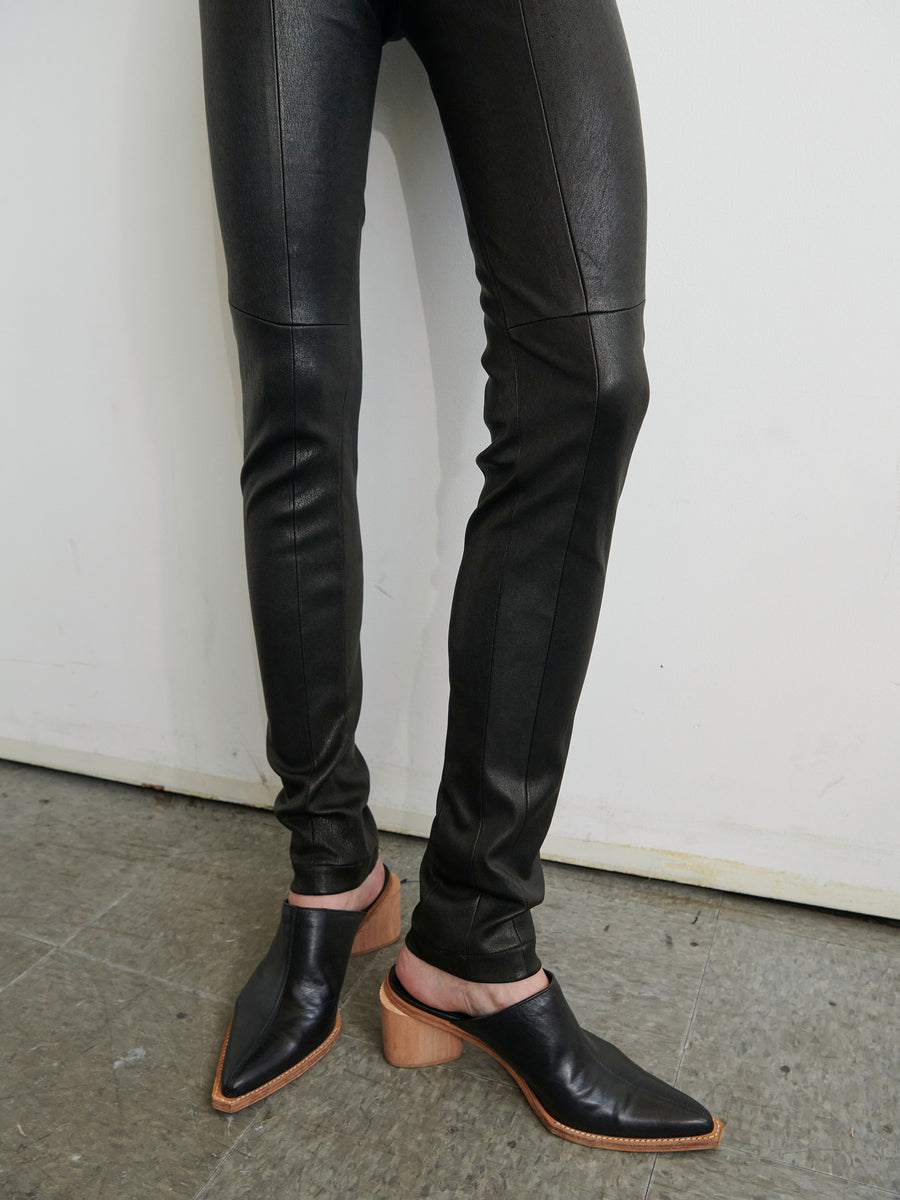 A person wearing Zero + Maria Cornejo's Leather One Seam Pant with an elastic waistband and black pointed-toe shoes featuring wooden block heels stands on a grey tiled floor against a plain white wall. The image focuses on their legs and footwear.