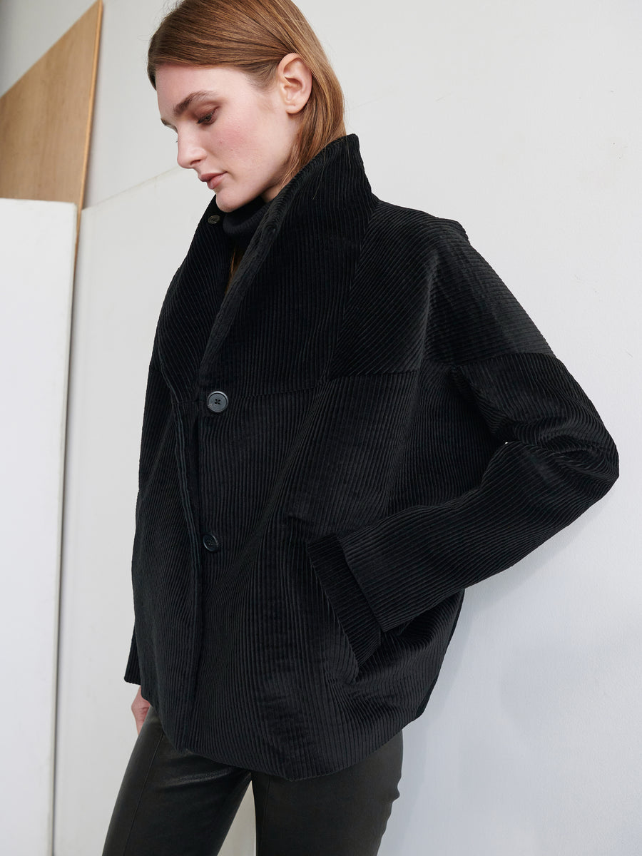 A person with long hair wearing a loose-fitting Osita Bomber jacket from Zero + Maria Cornejo, characterized by its black cotton corduroy fabric and large buttons, stands against a minimalist white and beige background, looking downward with one hand in their pocket.