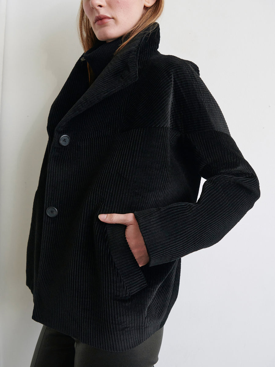 A person is wearing the Osita Bomber by Zero + Maria Cornejo, a black, wide-wale corduroy jacket with two large buttons and their right hand in the pocket, standing against a plain white background. The jacket features a slightly oversized fit and high collar.