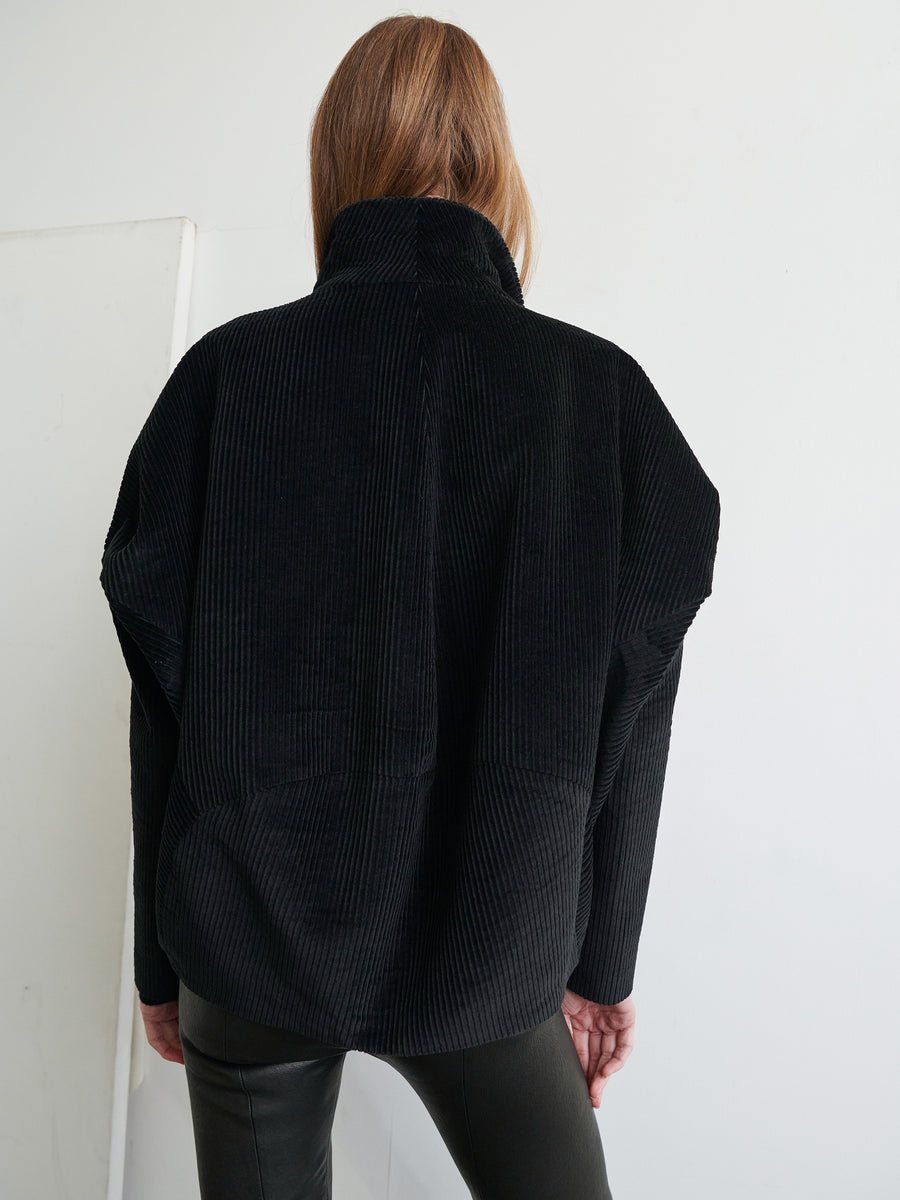 A person with long brown hair stands facing a white wall, wearing the Osita Bomber by Zero + Maria Cornejo paired with black pants. The wide-wale cotton corduroy jacket features a high collar and loose fit. The person's left arm is slightly bent, and their hand is partially visible.