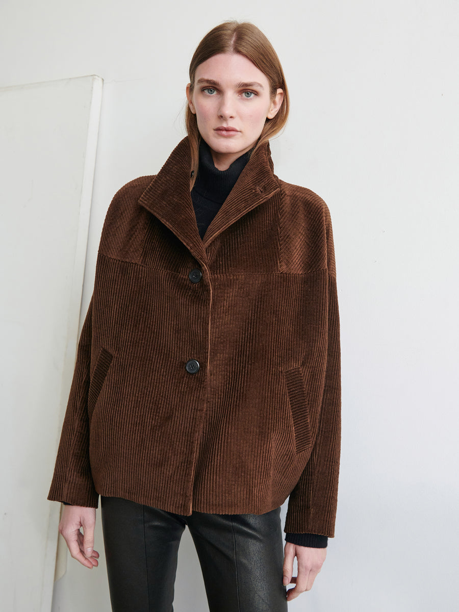 A person with long hair, wearing the Zero + Maria Cornejo Osita Bomber— a brown cotton wide-wale corduroy jacket with large buttons and a high collar— stands against a plain white background. The individual has one hand partially in a pocket, looking directly at the camera.