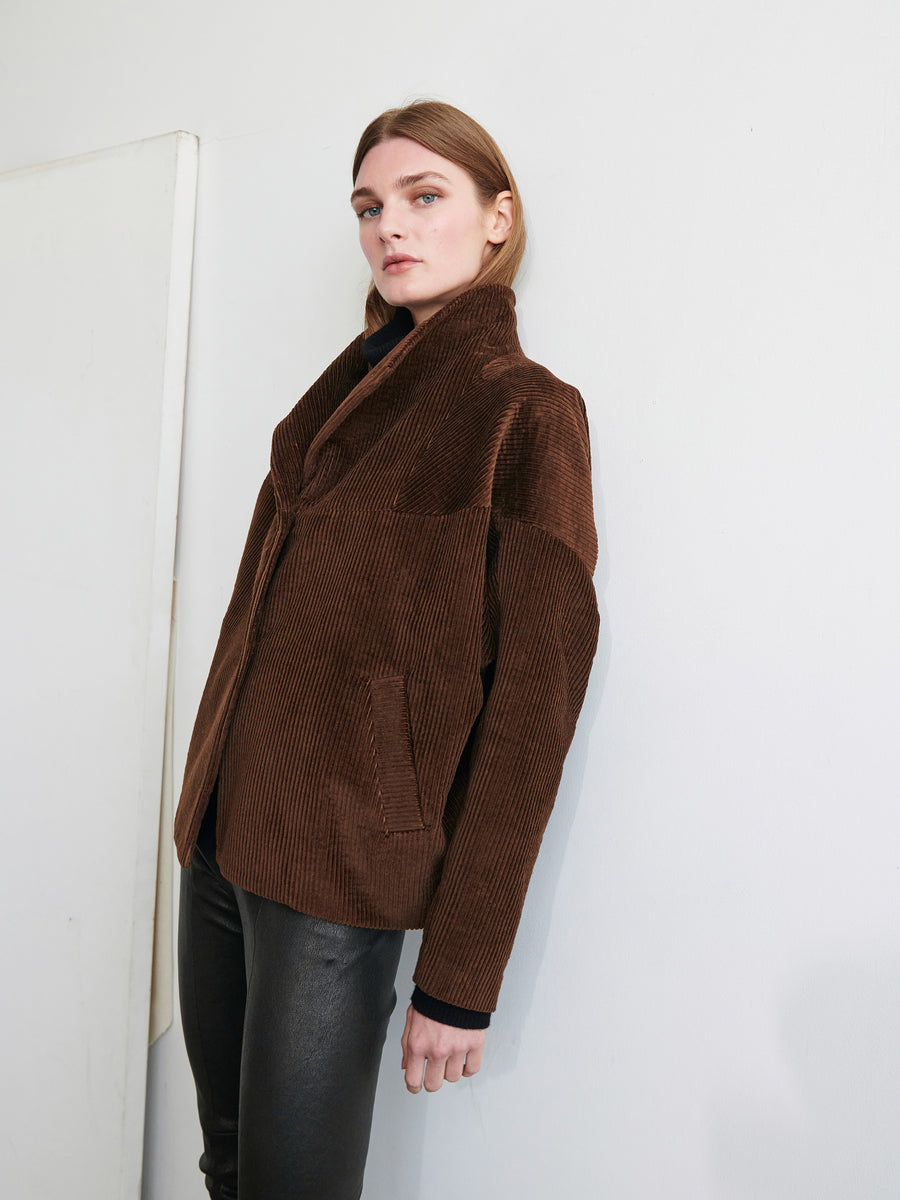 A person leans against a wall wearing the Osita Bomber, a brown cotton corduroy jacket with a high collar from Zero + Maria Cornejo, paired with black pants. They have long, straight hair and look directly at the camera with a neutral expression against a plain and minimalistic background.