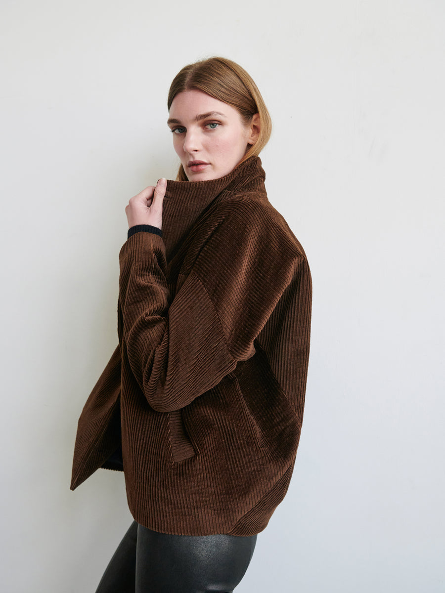 A person with medium-length, straight brown hair is wearing the Osita Bomber by Zero + Maria Cornejo paired with black leather pants. Made in New York, they are looking at the camera with a neutral expression while holding the collar of their high-collared, dark brown wide-wale corduroy jacket with one hand. The background is plain white.