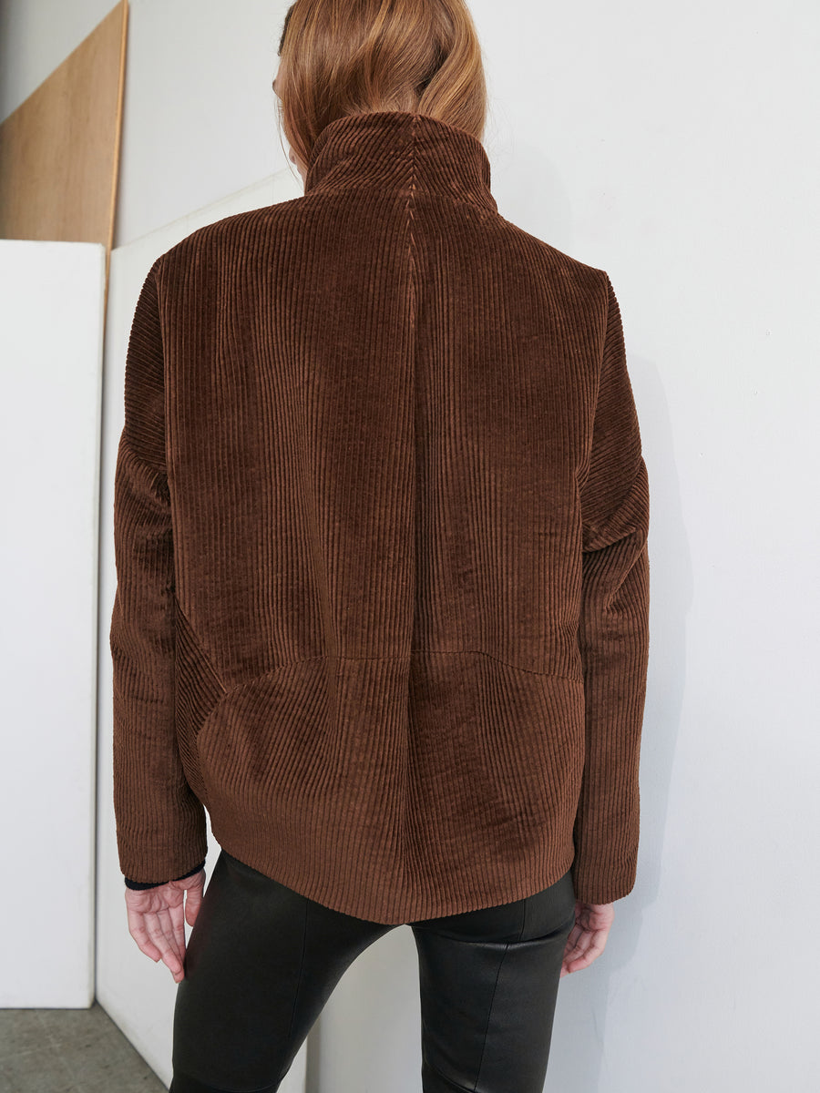 A person with shoulder-length hair is standing with their back to the camera, wearing a high-collared Osita Bomber jacket in brown wide-wale corduroy from Zero + Maria Cornejo and black pants. The background shows a white wall and a partial view of a wooden panel.