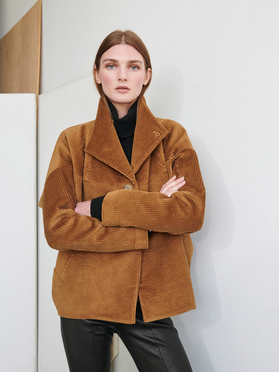 A woman with straight brown hair, dressed in a black turtleneck and the Osita Bomber by Zero + Maria Cornejo, stands with her arms crossed, gazing directly at the camera. The background features white and wooden surfaces.