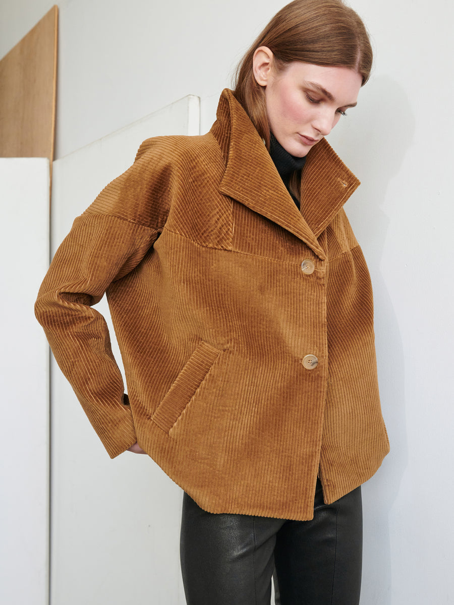 A person stands indoors wearing a Zero + Maria Cornejo Osita Bomber, featuring wide lapels and large buttons. They are looking down, hands resting on their hips, dressed in black pants. The background showcases a light-colored wall and door, subtly emphasizing the sophistication of their Made in New York attire.