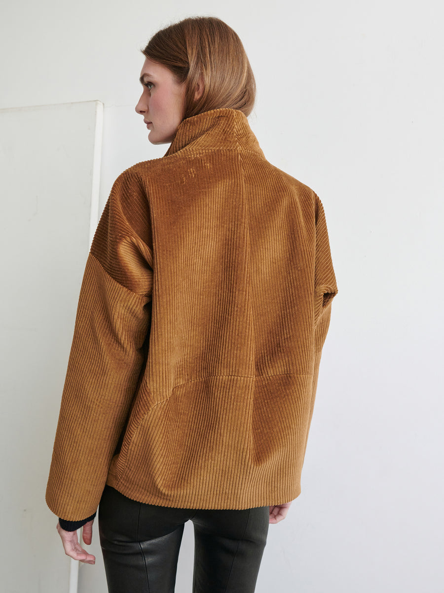 A person with long light brown hair is seen from the back, wearing a tan Zero + Maria Cornejo Osita Bomber jacket with a high collar. The person is also wearing black pants and standing against a plain white background. The stylish wide-wale design of the jacket adds a touch of classic elegance to the look.