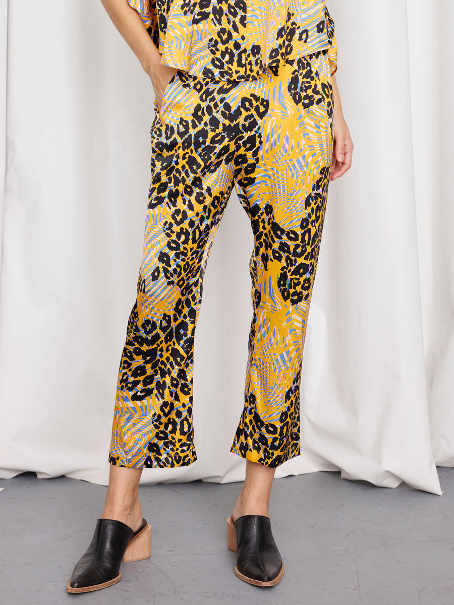 A person stands with hands in their pockets, wearing Zero + Maria Cornejo's Eko Pant—abstract animal print silk trousers with an elastic waist—paired with black slip-on shoes, set against a white curtain and gray floor.