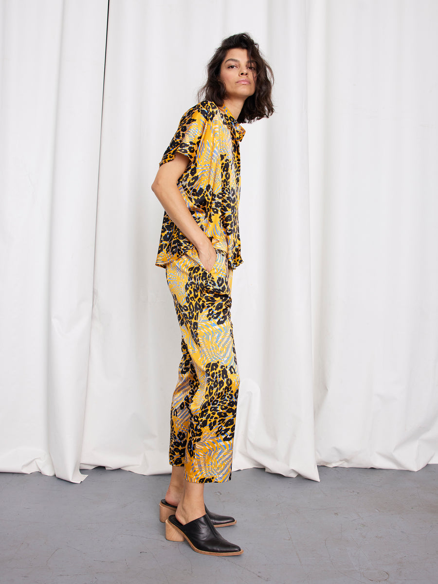 A person with shoulder-length dark hair stands sideways in front of white curtains, wearing Zero + Maria Cornejo Eko Pant and matching abstract animal print silk shirt, with black heeled mules and hands in pockets.