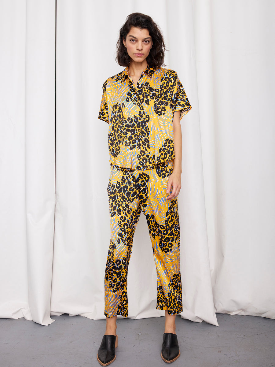 A person stands before white curtains wearing the Zero + Maria Cornejo Circle Shirt in a yellow, black, and light purple abstract animal print set with black slip-on shoes.