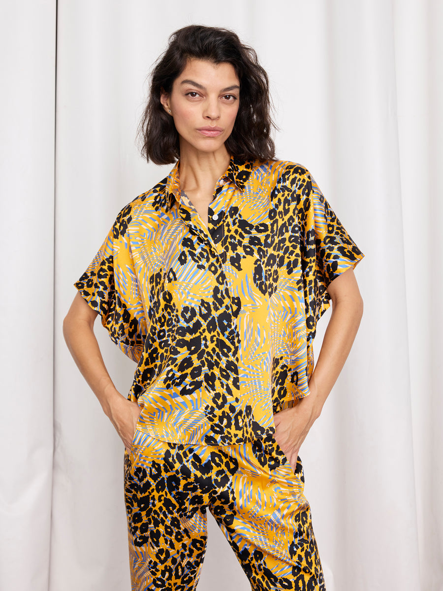 A woman with wavy dark hair stands with her hands in her pockets, wearing the Circle Shirt and matching pants from Zero + Maria Cornejo in a bold yellow, black, and blue print, made in New York, in front of white curtains.