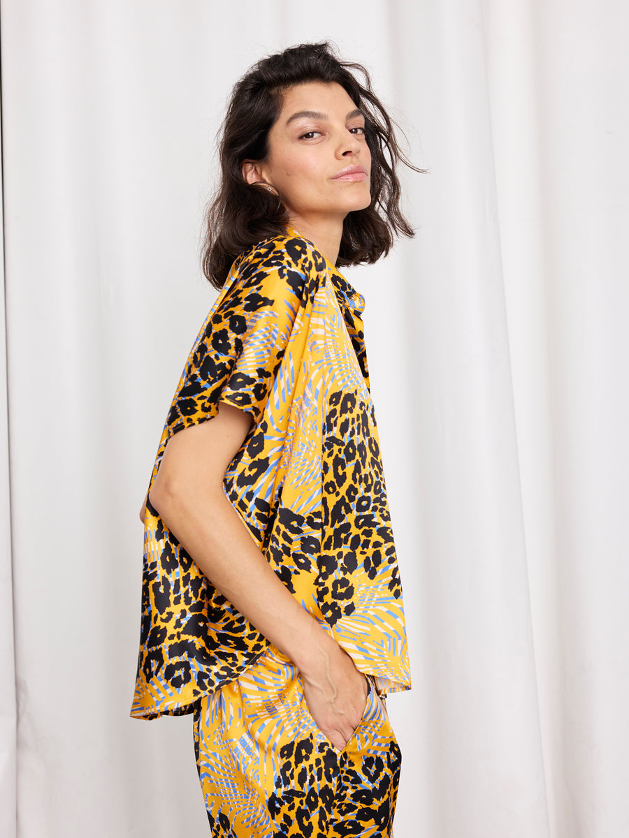 A person with medium-length dark hair wears the Zero + Maria Cornejo Circle Shirt, a loose-fitting silk short-sleeve top in yellow and black abstract animal print, paired with matching pants, standing sideways against a white curtain background.