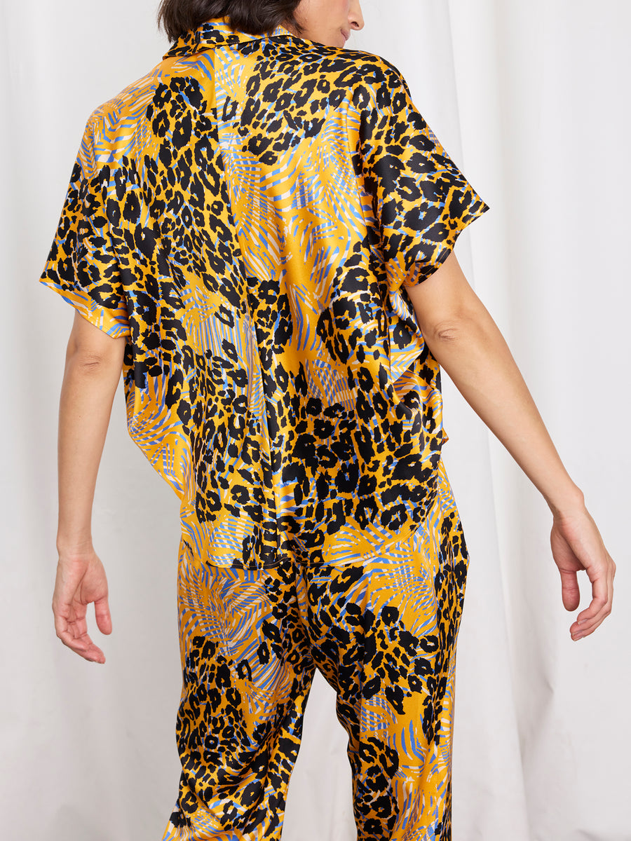 A person faces away from the camera, wearing the Zero + Maria Cornejo Circle Shirt—a silk short-sleeved top with a bold yellow, black, and blue leopard print—against a plain white curtain backdrop.