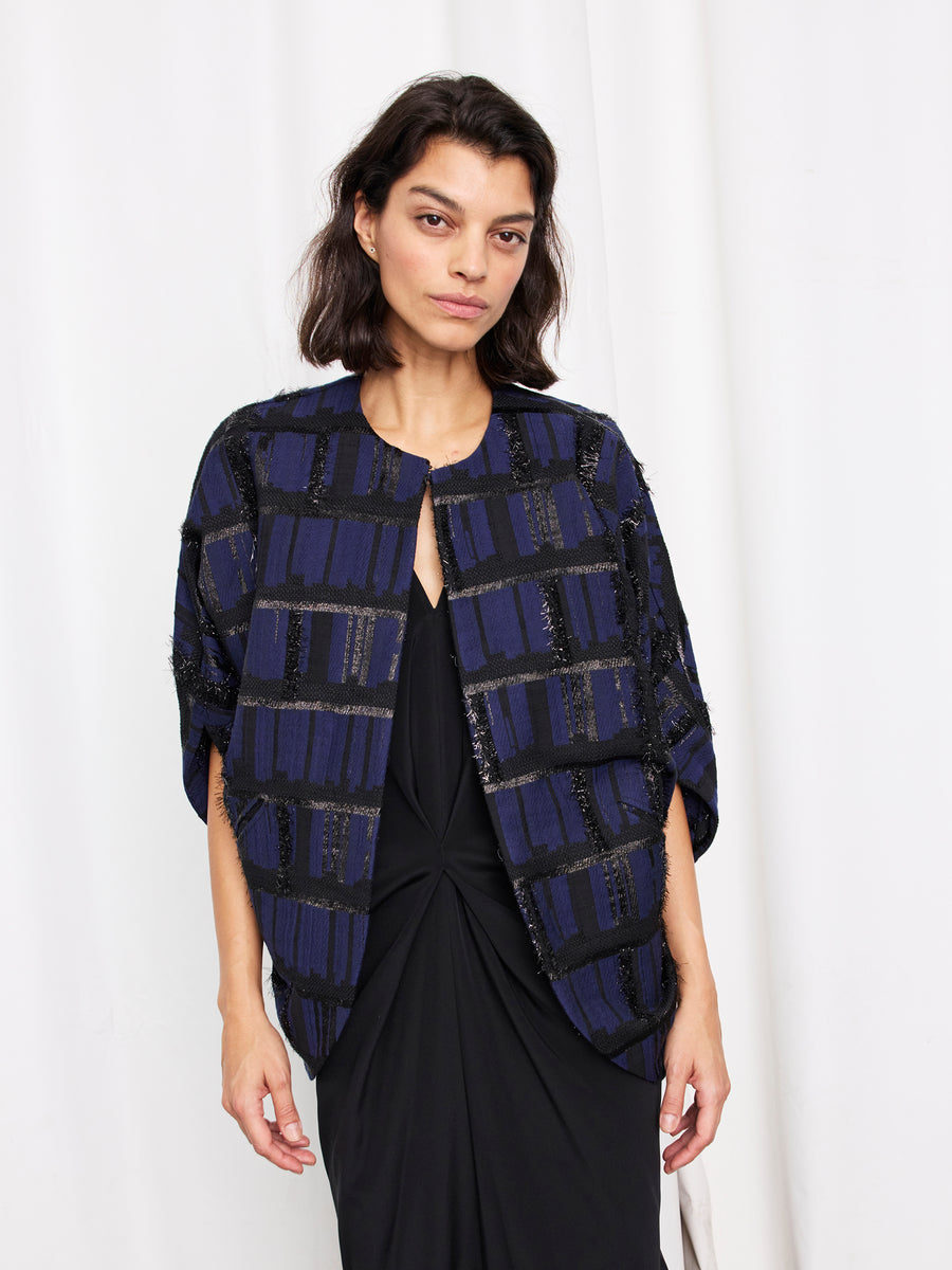 A woman with shoulder-length dark hair wears the Zero + Maria Cornejo Rio Shrug, a navy blue and black plaid jacket, over a black knotted-front dress, standing against a white curtain background.
