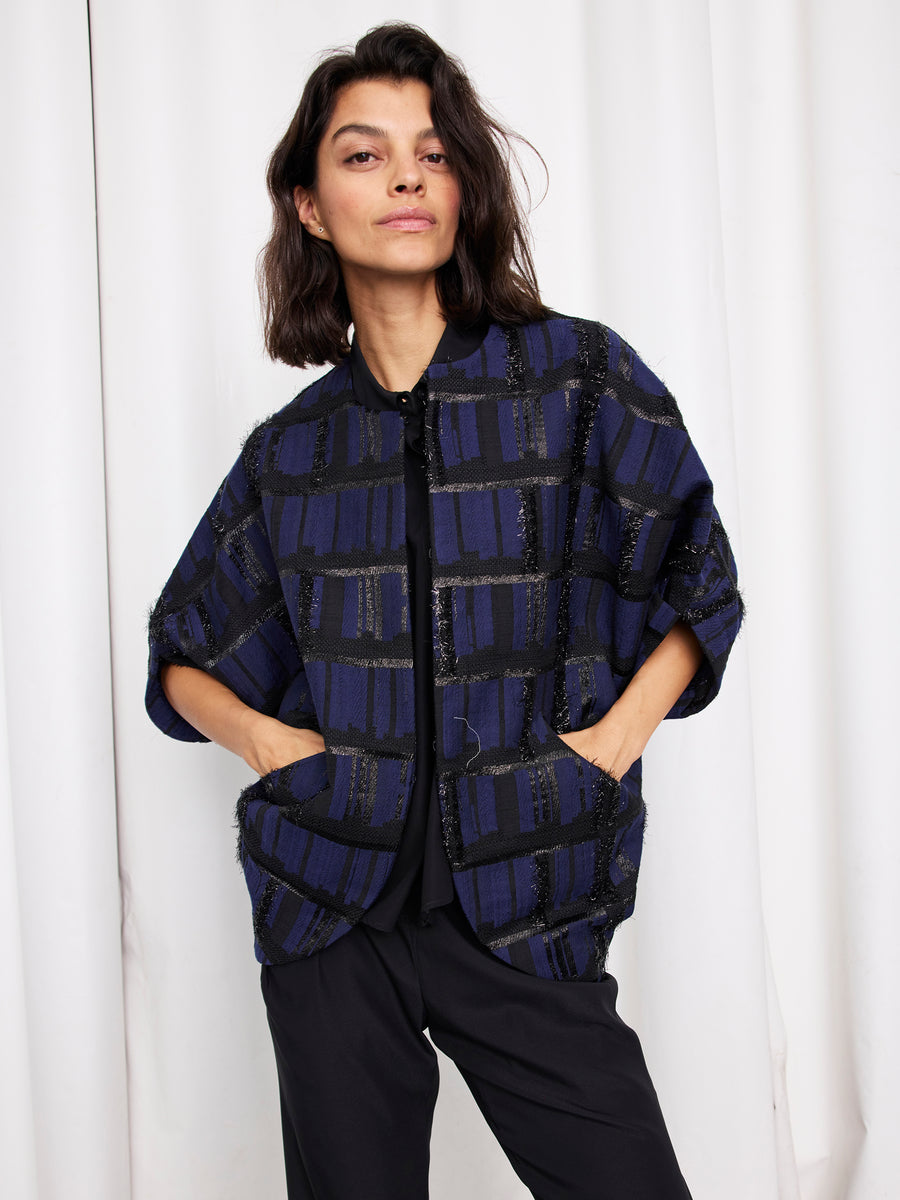 A woman stands confidently in front of a white curtain, wearing the Zero + Maria Cornejo Rio Shrug—a loose-fitting blue and black draped jacket—over black clothing, with her hands in her pockets and a slight smile.