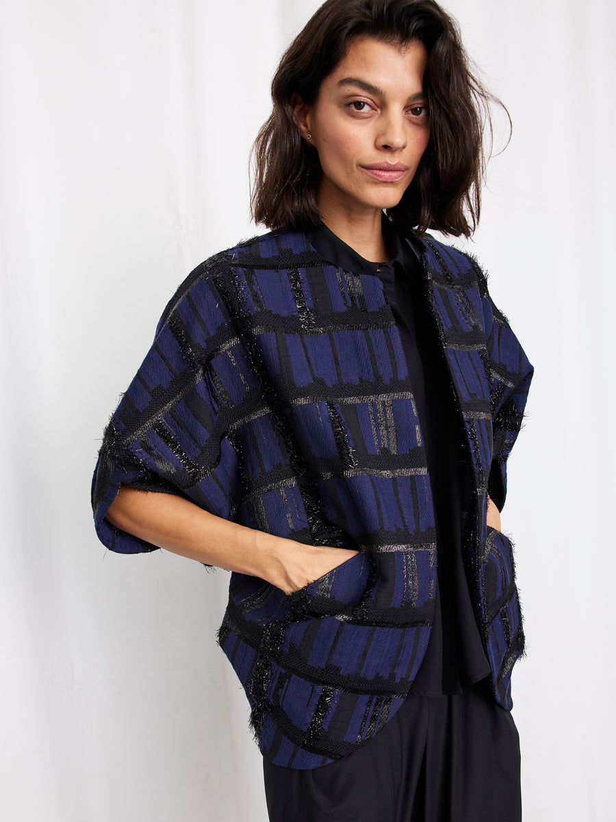 A woman with shoulder-length dark hair models the Zero + Maria Cornejo Rio Shrug, a blue and black plaid short-sleeved jacket, over a black top and pants, posing with her hands in the shrug's pockets against a white background.