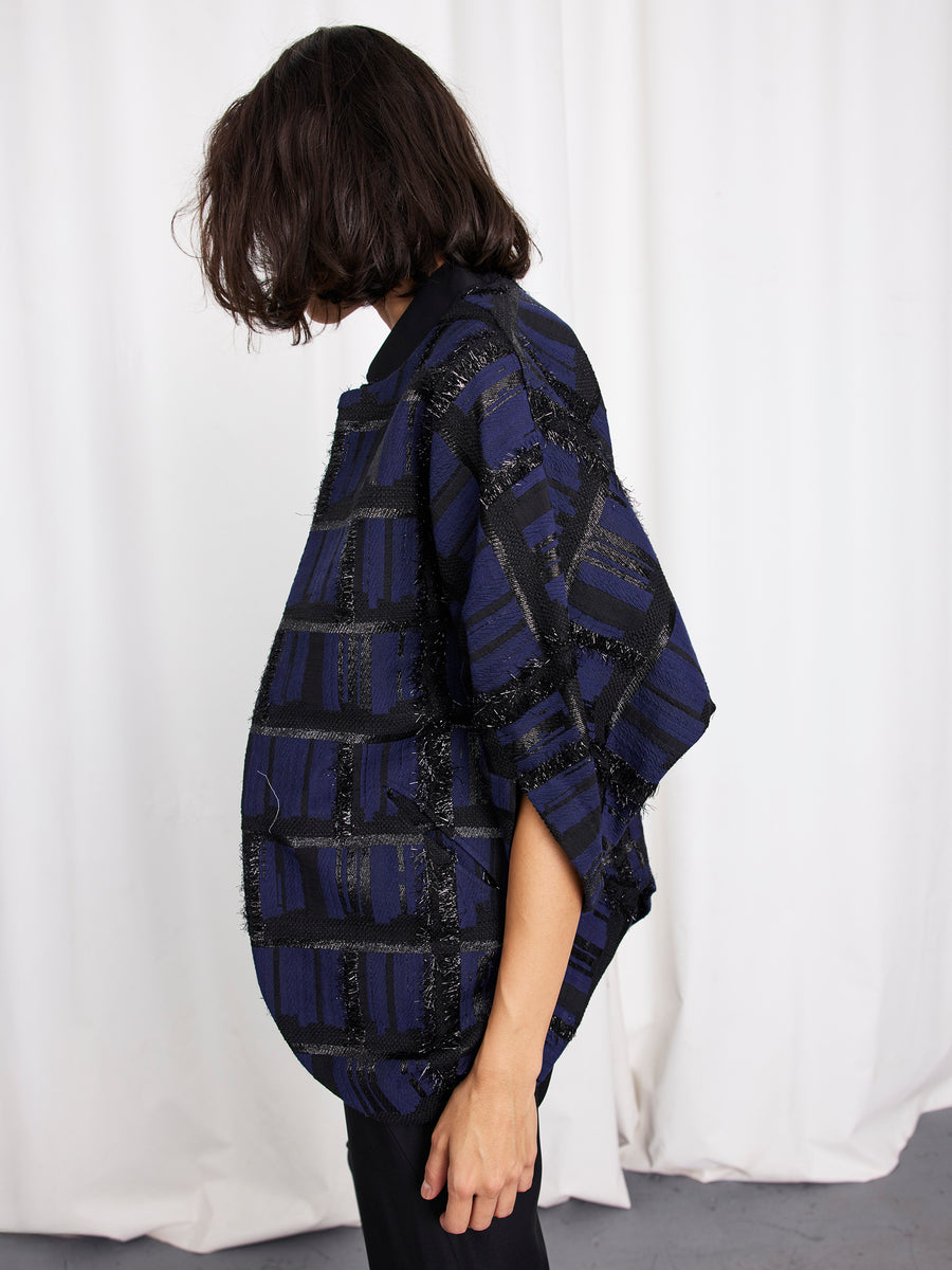 A person with shoulder-length dark hair, facing sideways, wears the Zero + Maria Cornejo Rio Shrug—an oversized dark blue and black plaid jacket with textured details. White curtains hang in the background.