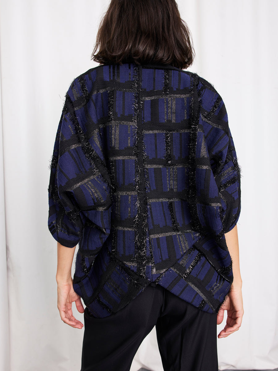 A person with shoulder-length dark hair, seen from behind, wears the Rio Shrug by Zero + Maria Cornejo—a loose-fitting metallic jacquard jacket in navy and black checks—paired with black pants, standing before a white curtain.
