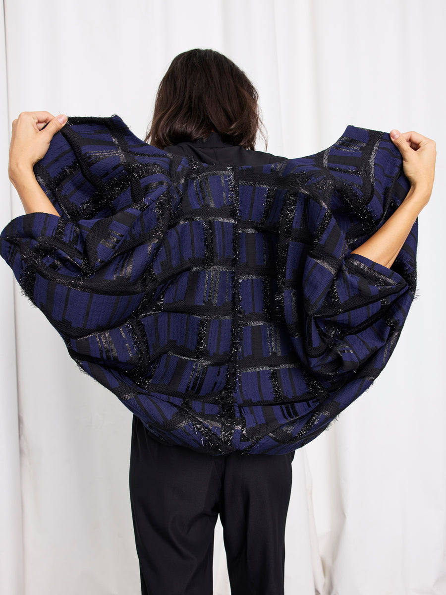 A person stands facing away, holding up the back of the Zero + Maria Cornejo Rio Shrug in dark blue and black metallic jacquard, showing off its wide, textured fabric against a white curtain backdrop.