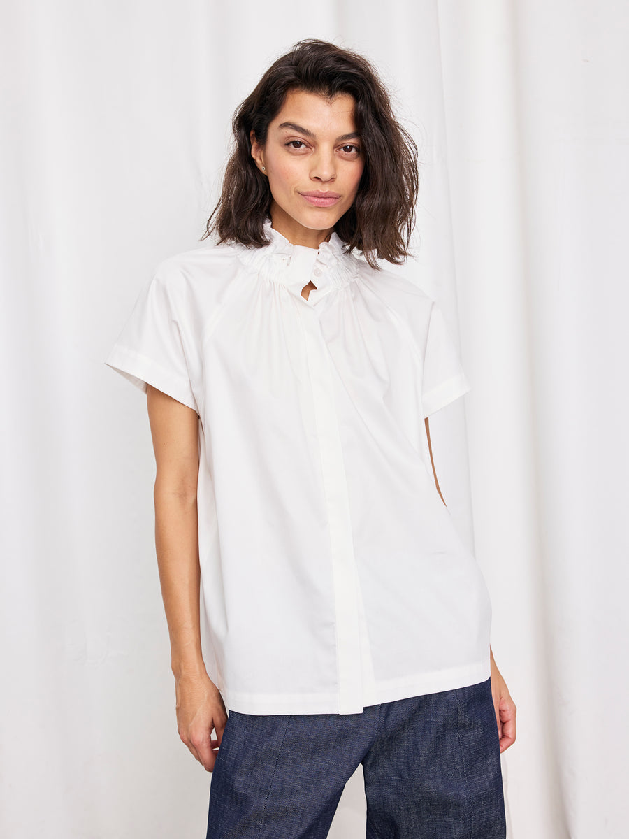 A person with shoulder-length dark hair wears the Zero + Maria Cornejo Elastic Gaban Shirt, featuring a ruffle collar, styled with dark blue pants against a white curtain backdrop.