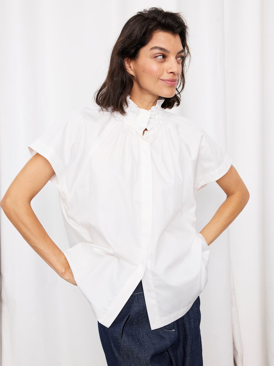 A woman with dark shoulder-length hair stands against a white curtain, wearing the Zero + Maria Cornejo Elastic Gaban Shirt and dark blue pants, hands on her hips and a slight smile on her face.