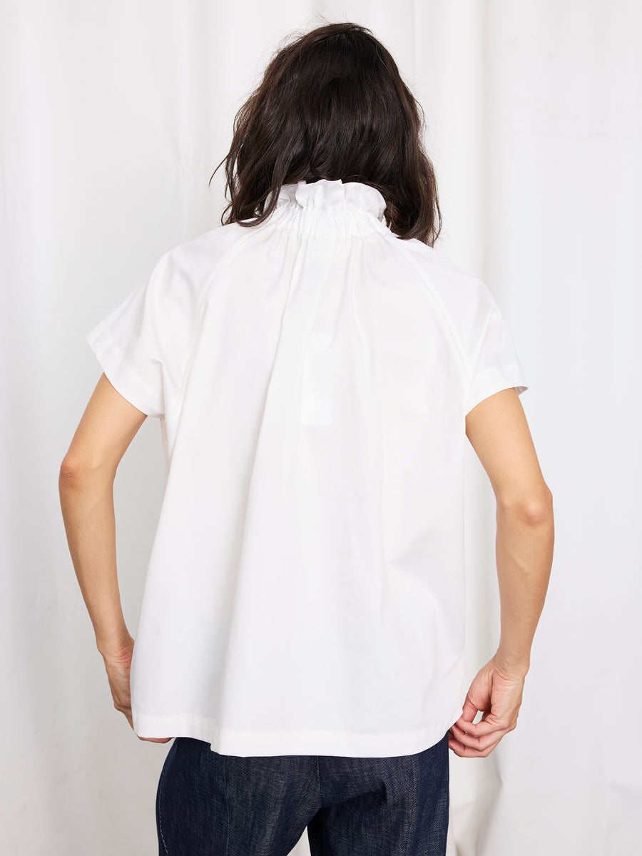 A person with shoulder-length dark hair wearing the Zero + Maria Cornejo Elastic Gaban Shirt—white with a ruffle collar—seen from the back, paired with dark pants against a light background. Made in New York.