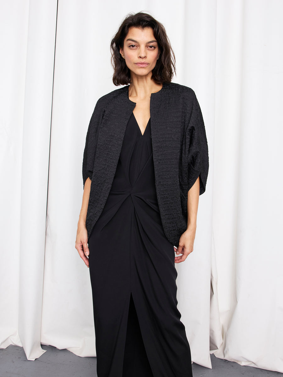 A woman with shoulder-length dark hair stands before white curtains, wearing the Zero + Maria Cornejo Rio Shrug—crafted from recycled polyester—over a black dress with knot detail and front slit, gazing calmly at the camera.