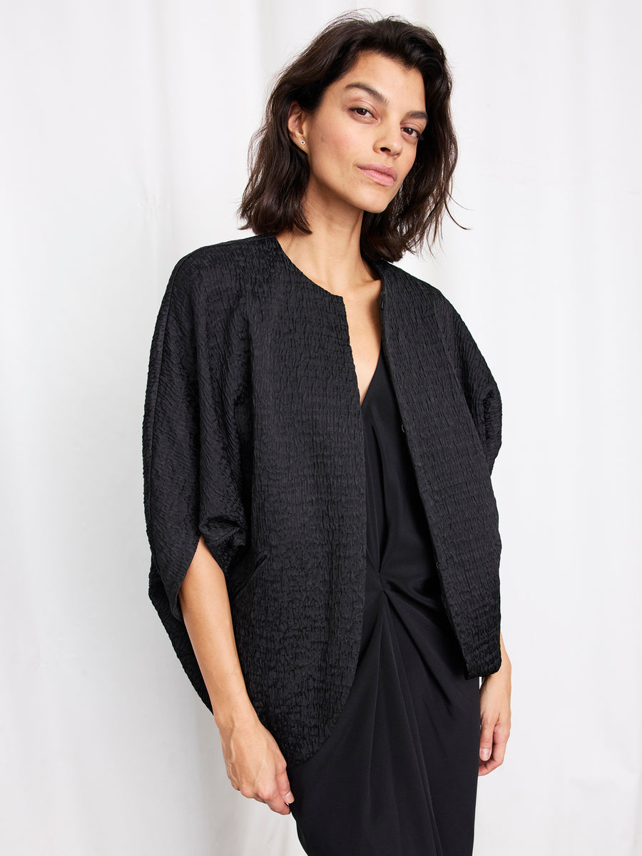 A woman with shoulder-length dark hair wears the Rio Shrug by Zero + Maria Cornejo, a crinkle-effect jacket crafted from recycled polyester, layered over a black dress. She stands against a plain white background facing the camera neutrally.