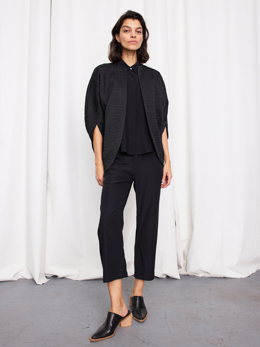 A woman with short dark hair stands against white curtains, hands in her pockets, wearing the Rio Shrug by Zero + Maria Cornejo—a loose black crinkle-effect jacket made from recycled polyester—paired with black pants and mule shoes.